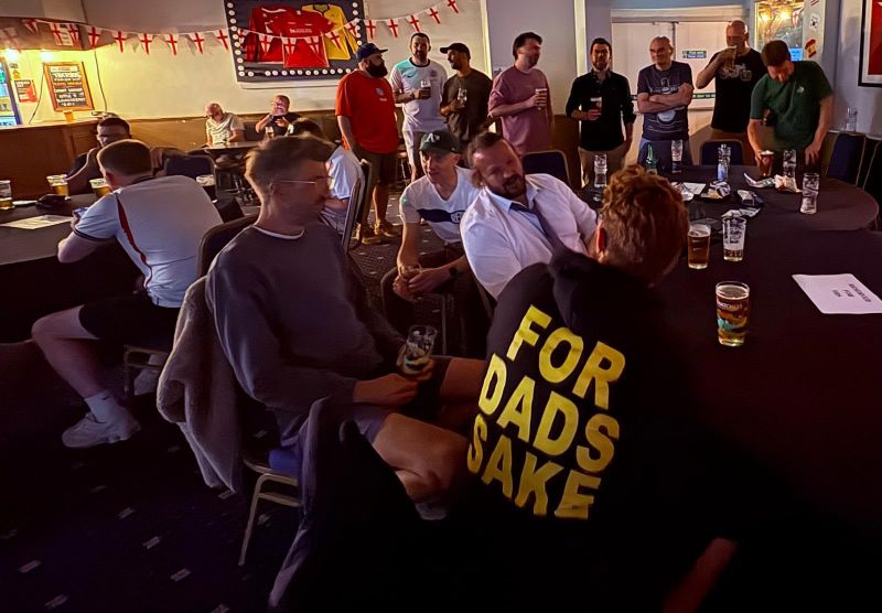 A packed room of dads watching the football, one in a For Dads Sake hoodie