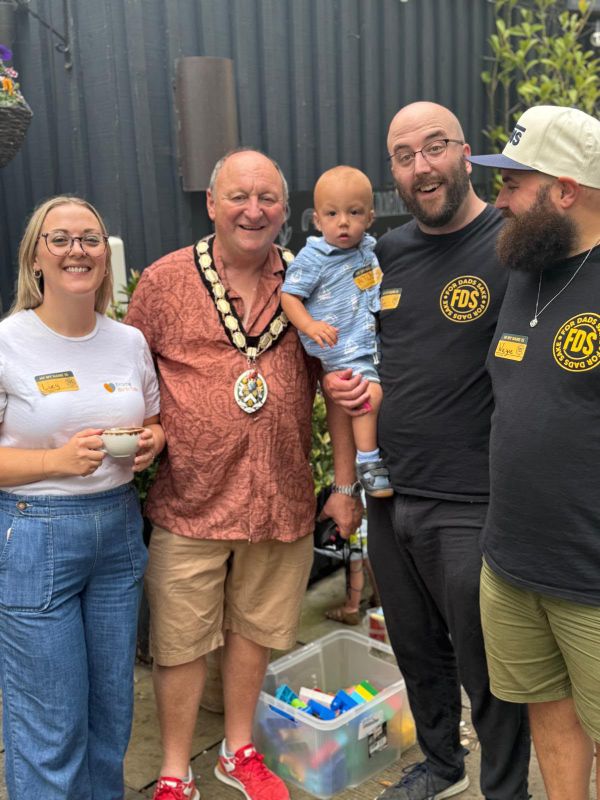 A group photo with a guest wearing a chain of office, a toddler and two dads in FDS t-shirts