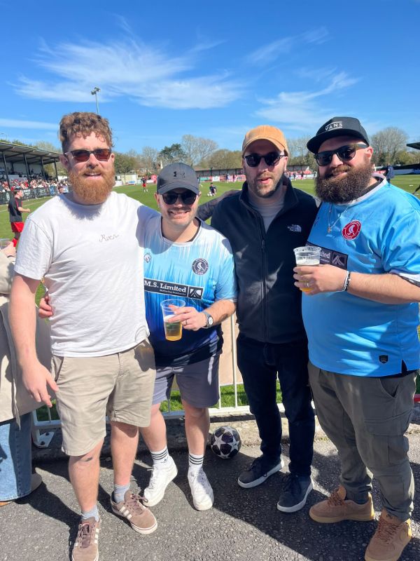Four dads with drinks at the side of a football pitch on a sunny day