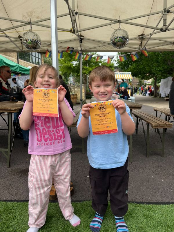 Two children holding FDS anniversary party flyers under a gazebo